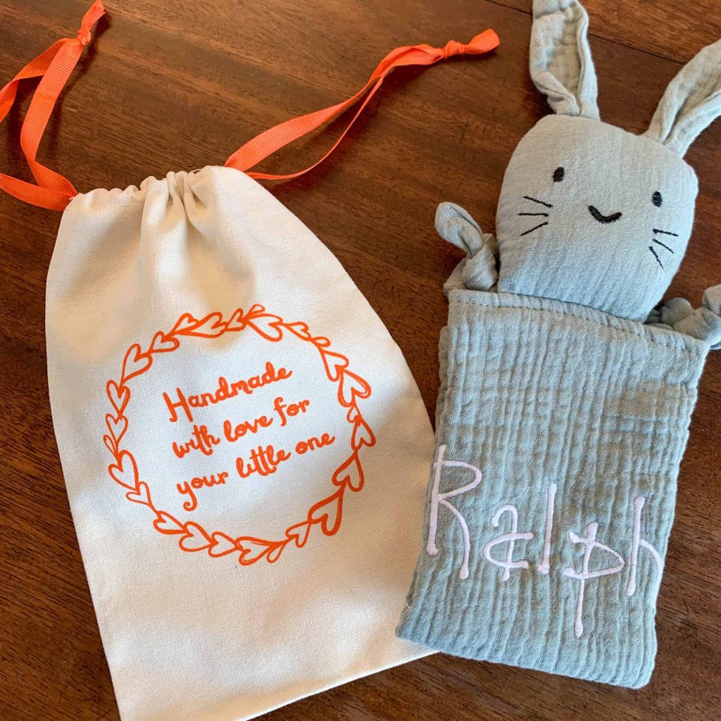 A handmade sage green muslin bunny comforter embroidered with the name 'Ralph' next to a cream drawstring gift bag