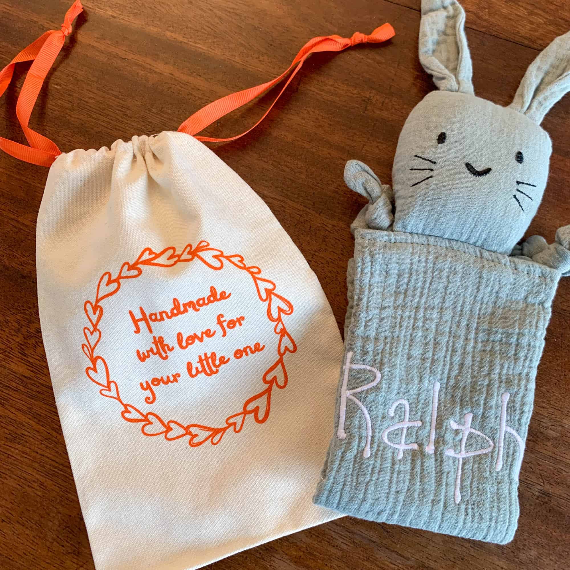 A handmade sage green muslin bunny comforter embroidered with the name 'Ralph' next to a cream drawstring gift bag