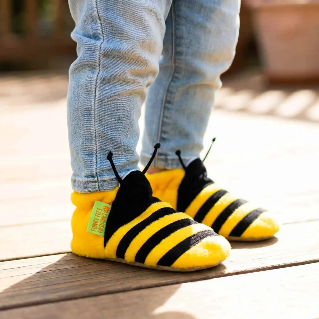 Child in Funky Feet Fashions bumble bee booties with yellow stripes and antennae, standing on a wooden deck.