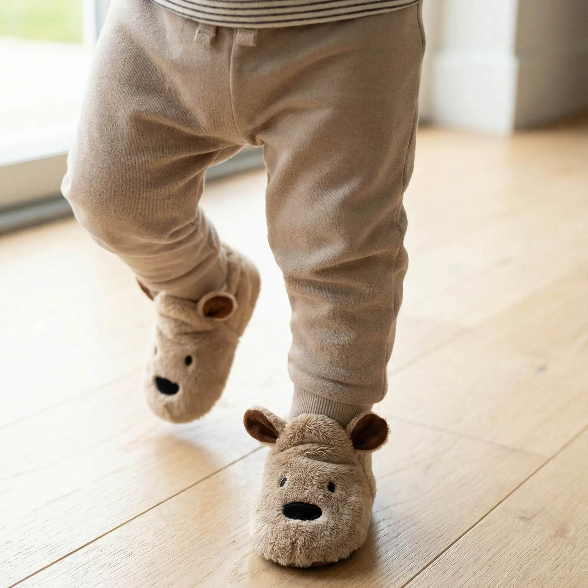 Toddler in brown bear baby booties by Funky Feet Fashions, walking in tan joggers on a light wooden floor.